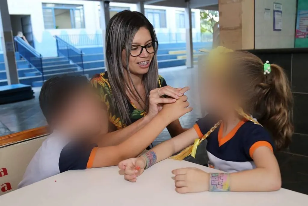 Aline sentada à mesa com duas crianças no pátio da escola (rostos desfocados)